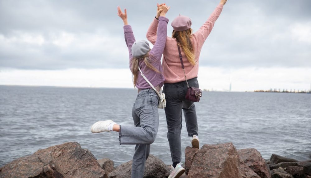 Two women in hats joyfully pose on seaside rocks, enjoying a cloudy day.