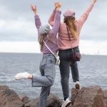 Two women in hats joyfully pose on seaside rocks, enjoying a cloudy day.