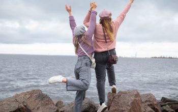 Two women in hats joyfully pose on seaside rocks, enjoying a cloudy day.